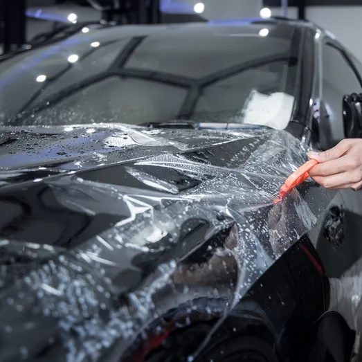 Technician applying clear paint protection film to a black car hood with a squeegee